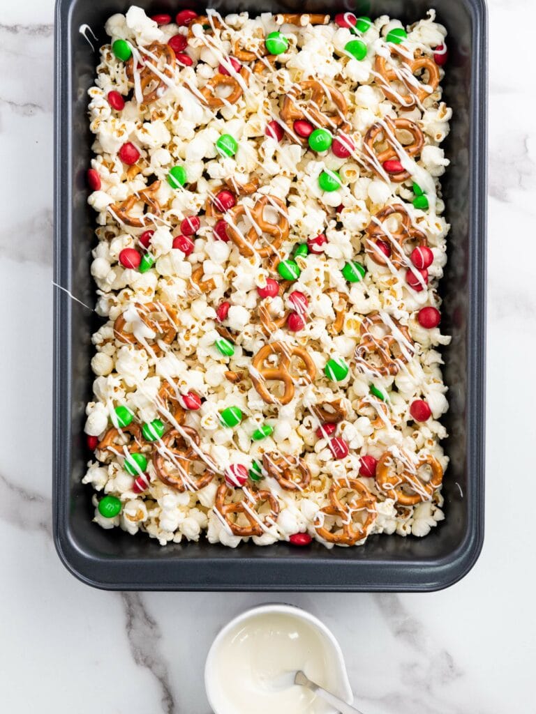 A baking pan filled with popcorn, pretzels, red and green candy-coated chocolates, all drizzled with white chocolate.
