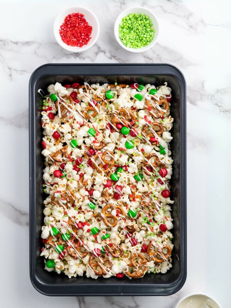A baking tray filled with popcorn, pretzels, red and green candies, and sprinkles, drizzled with white chocolate. Two small bowls of red and green sprinkles are nearby.