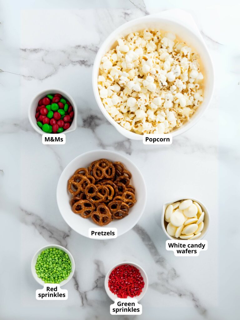 A bowl of popcorn surrounded by smaller bowls containing M&Ms, pretzels, white candy wafers, red sprinkles, and green sprinkles.