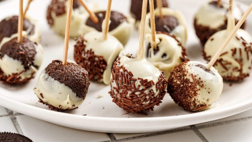 A plate of chocolate cake pops coated in white chocolate and decorated with chocolate sprinkles, each with a wooden stick inserted for easy handling.