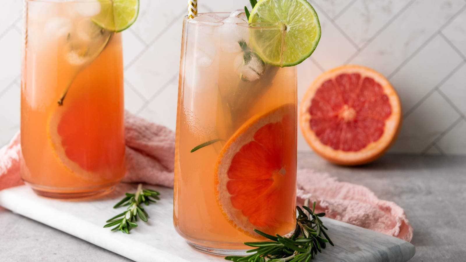 Two glasses of paloma cocktail with ice, garnished with rosemary and citrus slices.