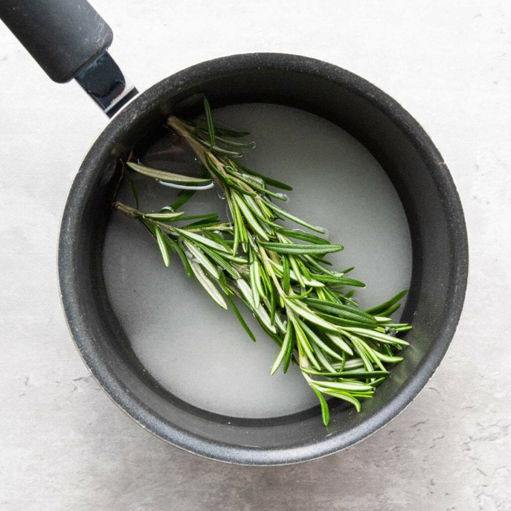 A small saucepan containing water, sugar, and fresh rosemary sprigs on a light gray surface.