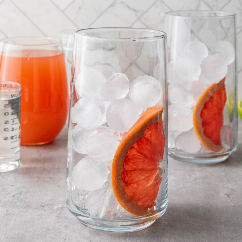 A glass filled with ice and a slice of grapefruit, with more glasses and a measuring cup in the background on a gray countertop.