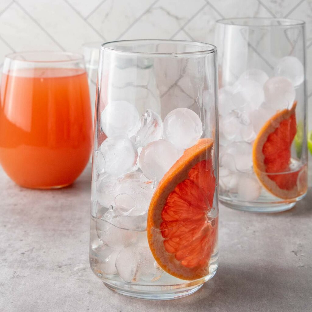 Two glasses with ice and a slice of grapefruit, next to a glass of grapefruit juice, on a gray surface with a tiled background.