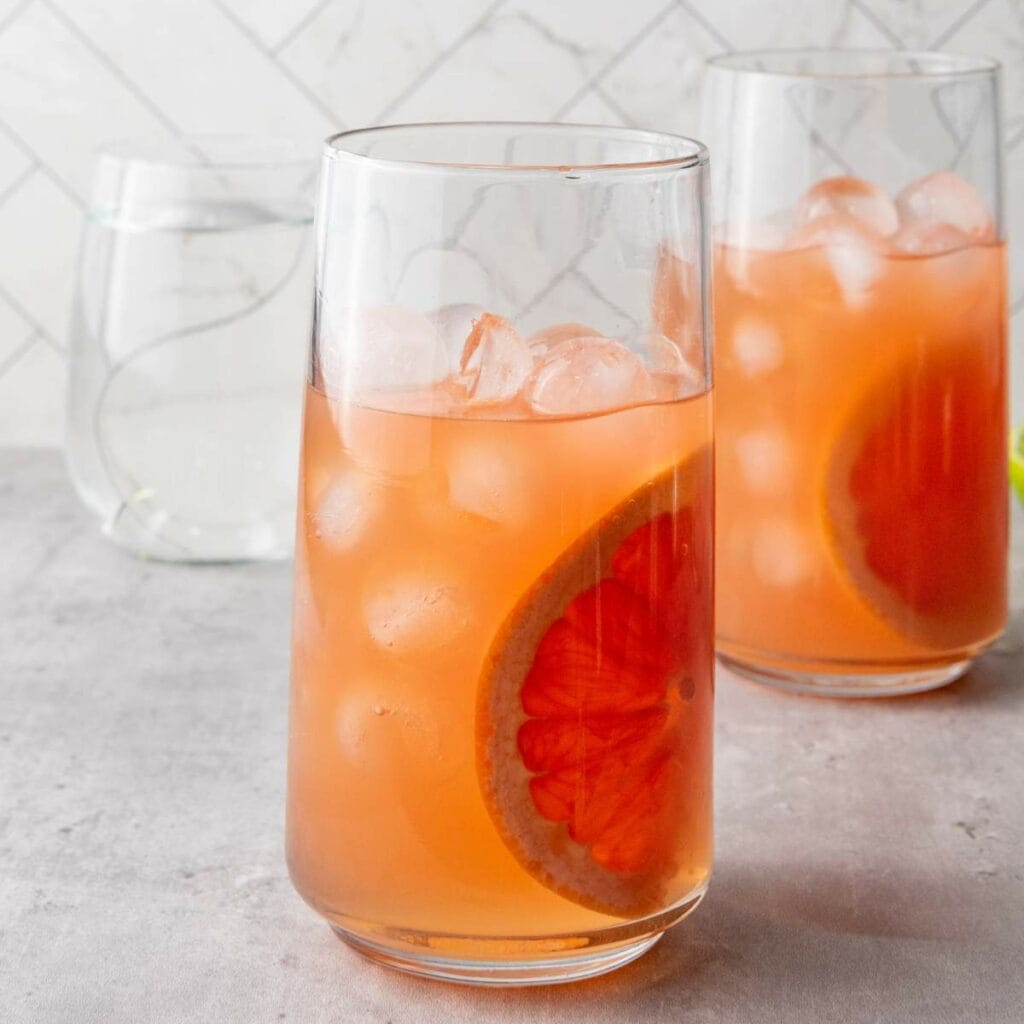 Two glasses filled with an orange-colored drink, ice cubes, and a slice of grapefruit, set on a gray surface.