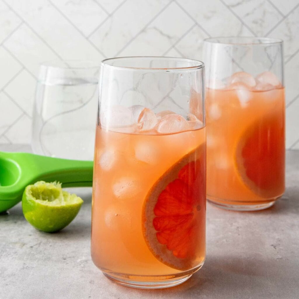 Two glasses of pink grapefruit drink with ice and grapefruit slice, a halved lime, and a green citrus squeezer on a gray countertop.