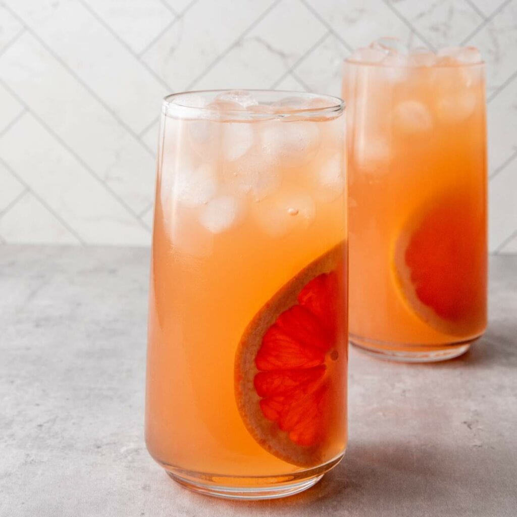 Two tall glasses filled with an orange-pink iced drink, each garnished with a slice of grapefruit.