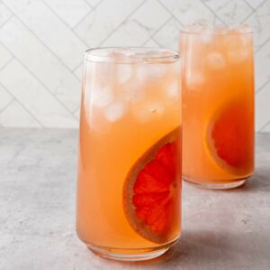 Two tall glasses filled with an orange-pink iced drink, each garnished with a slice of grapefruit.