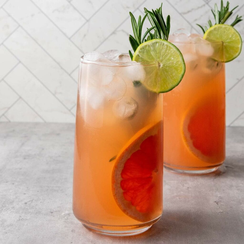 Two tall glasses of grapefruit cocktail with ice, garnished with lime slices and rosemary.