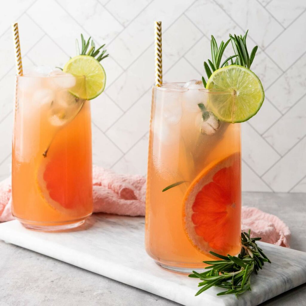 Two tall glasses of grapefruit cocktail with ice, lime slices, and rosemary sprigs on a marble surface.