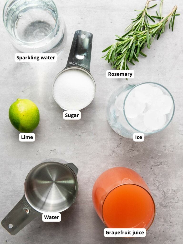 Labeled ingredients on a gray surface: sparkling water, rosemary, ice, grapefruit juice, water, sugar, and a lime.