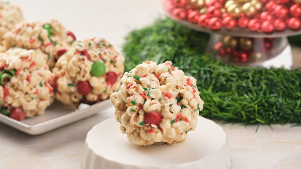 A Christmas popcorn ball with red and green candies and sprinkles sits on a white surface.