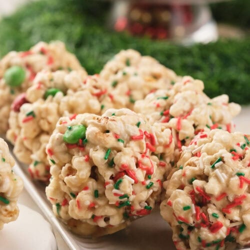 Festive popcorn balls with red and green sprinkles are arranged on a white plate, with a blurred festive background.
