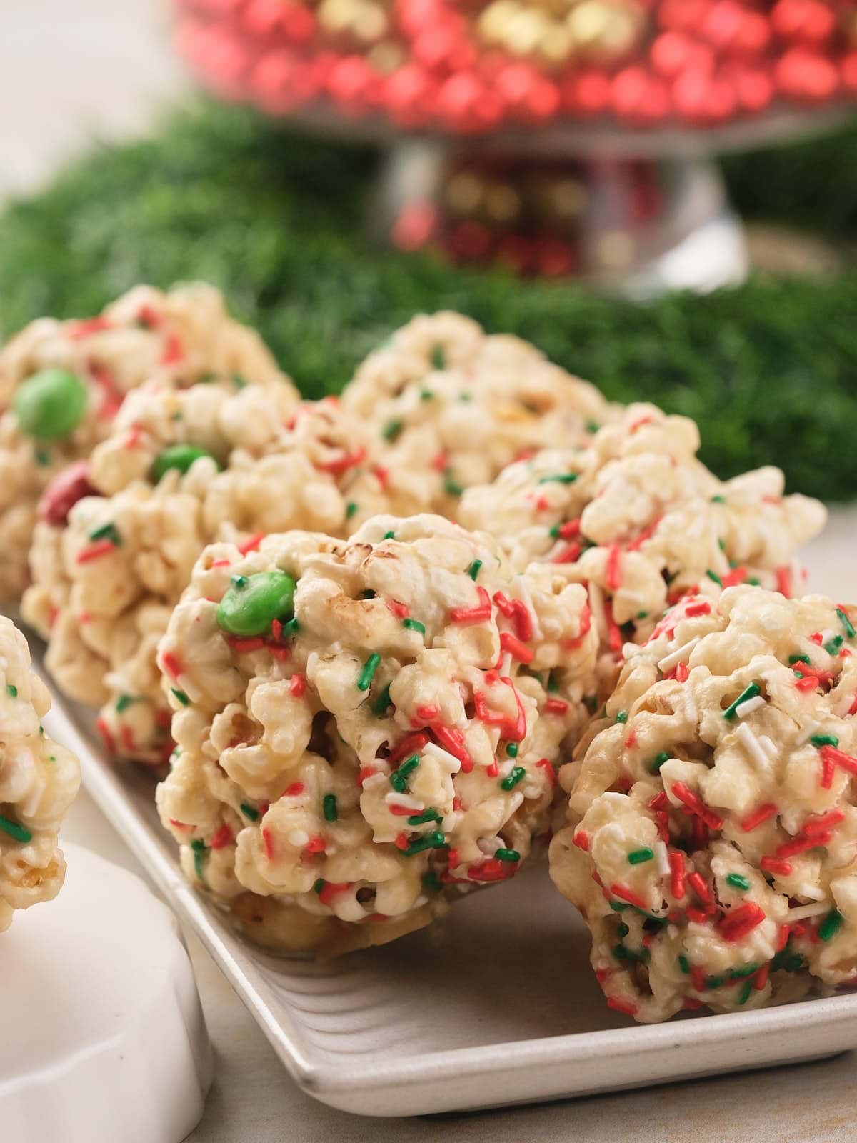 Festive popcorn balls with red and green sprinkles are arranged on a white plate, with a blurred festive background.