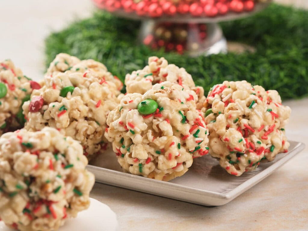 Popcorn balls mixed with red and green candy and sprinkles are arranged on a rectangular plate, with a festive centerpiece in the background.