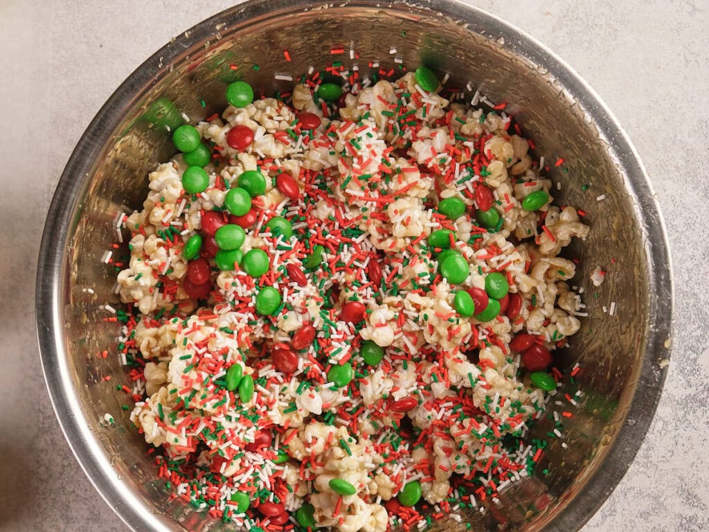 A metal bowl filled with popcorn mixed with red and green candy-coated chocolates and red, green, and white sprinkles.