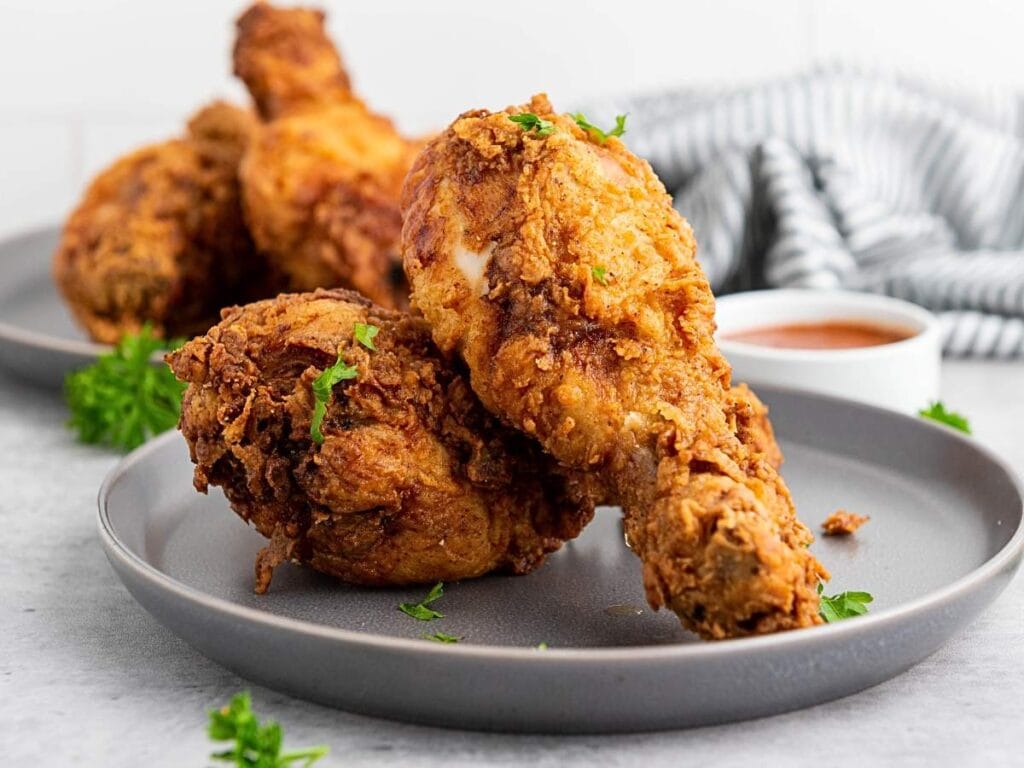 Two pieces of crispy fried chicken on a gray plate, garnished with parsley, with more chicken, a striped cloth, and a cup of sauce in the background.