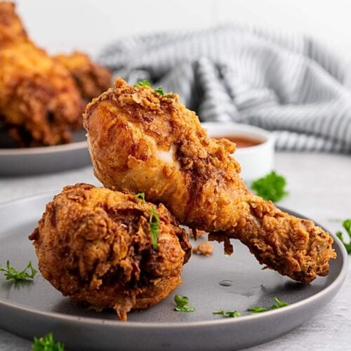 Two pieces of fried chicken, a drumstick and a thigh, are served on a gray plate with parsley garnish; more chicken and sauce appear in the background.