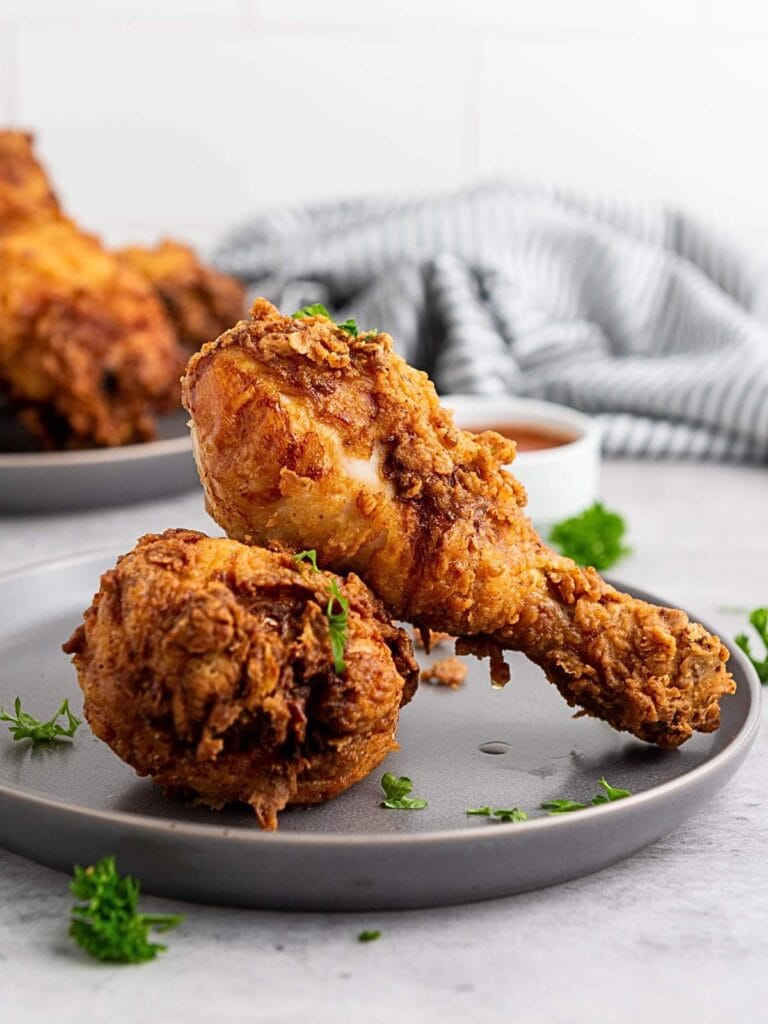 Two pieces of fried chicken, a drumstick and a thigh, are served on a gray plate with parsley garnish; more chicken and sauce appear in the background.
