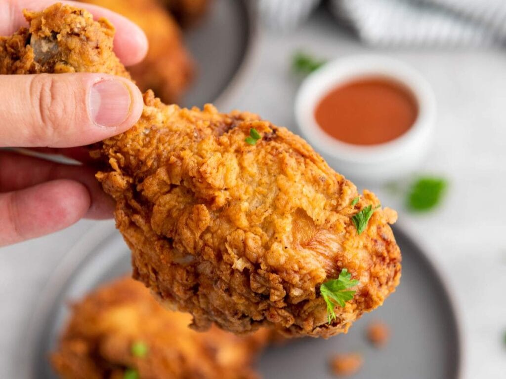 A hand holding a piece of crispy fried chicken drumstick garnished with parsley, with a plate of more chicken and a bowl of dipping sauce in the background.