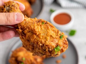 A hand holding a piece of crispy fried chicken drumstick garnished with parsley, with a plate of more chicken and a bowl of dipping sauce in the background.