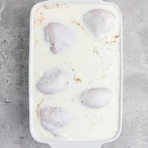 Raw chicken pieces are submerged in a white liquid marinade, likely buttermilk, inside a white rectangular baking dish on a gray countertop.