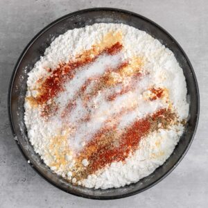A black bowl filled with flour, paprika, salt, pepper, and other seasonings on a light gray surface.