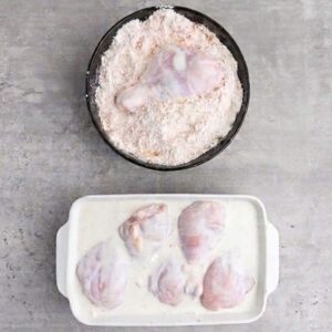 Raw chicken drumsticks and thighs are being coated in flour in a black bowl, with more chicken pieces marinating in a white liquid in a rectangular dish below.
