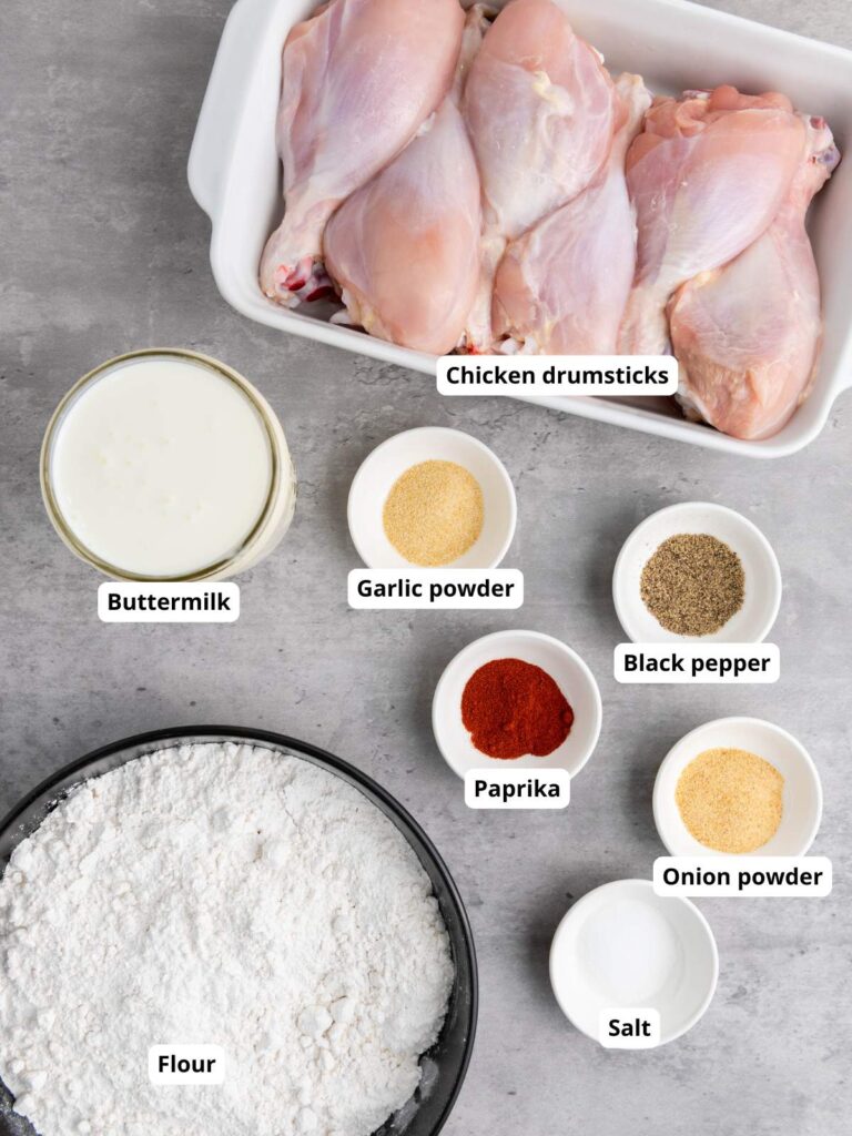 Raw chicken drumsticks in a dish, surrounded by bowls of buttermilk, flour, garlic powder, paprika, onion powder, salt, and black pepper.