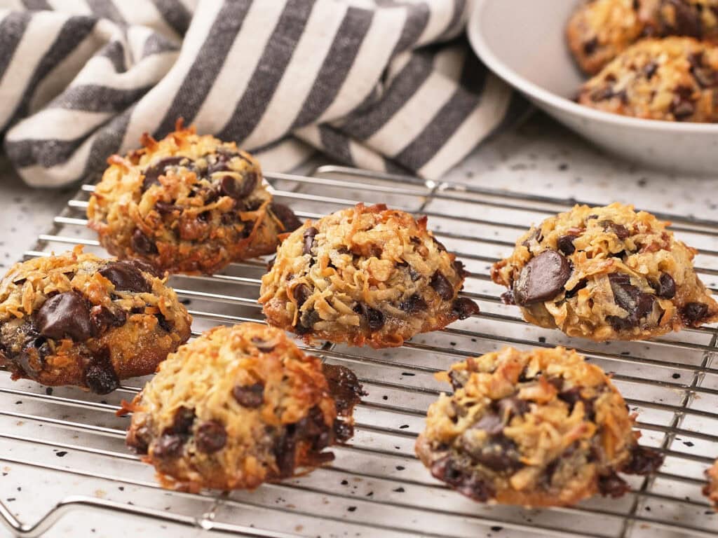 Seven magic cookies cooling on a wire rack, with a striped kitchen towel and a bowl of cookies in the background.