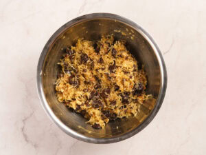 A metal mixing bowl containing a mixture of shredded coconut, raisins, and a sticky batter on a light-colored countertop.