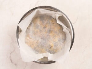A round metal bowl lined with parchment paper contains a dough mixture, viewed from above on a light-colored countertop.