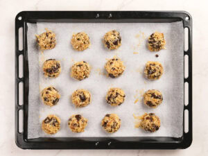A baking tray lined with parchment paper holds 16 evenly spaced balls of unbaked cookie dough with visible chocolate chips.