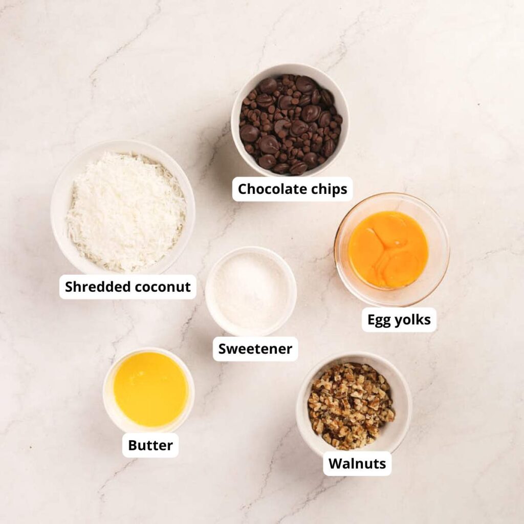 Top-down view of bowls containing chocolate chips, egg yolks, shredded coconut, sweetener, melted butter, and chopped walnuts.