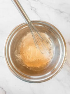 A metal mixing bowl containing flour and a metal whisk on a white marble surface.