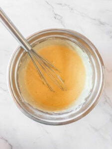 A metal whisk rests in a stainless steel bowl containing a smooth, light yellow batter on a white marble surface.