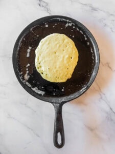 A pancake cooking in a black cast iron skillet on a white marble surface.
