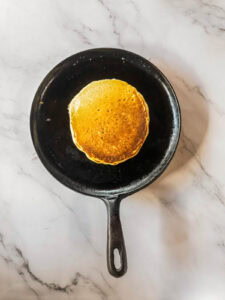 A single pancake cooking in a black cast iron skillet on a white marble surface.