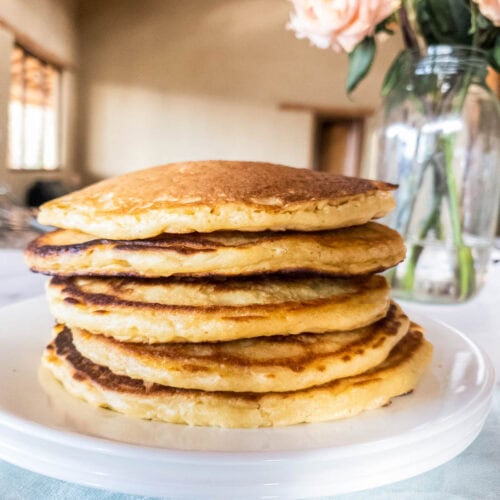 A stack of seven golden pancakes on a white plate with a glass vase of pink roses in the background.