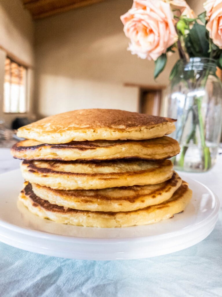 A stack of seven golden pancakes on a white plate with a glass vase of pink roses in the background.