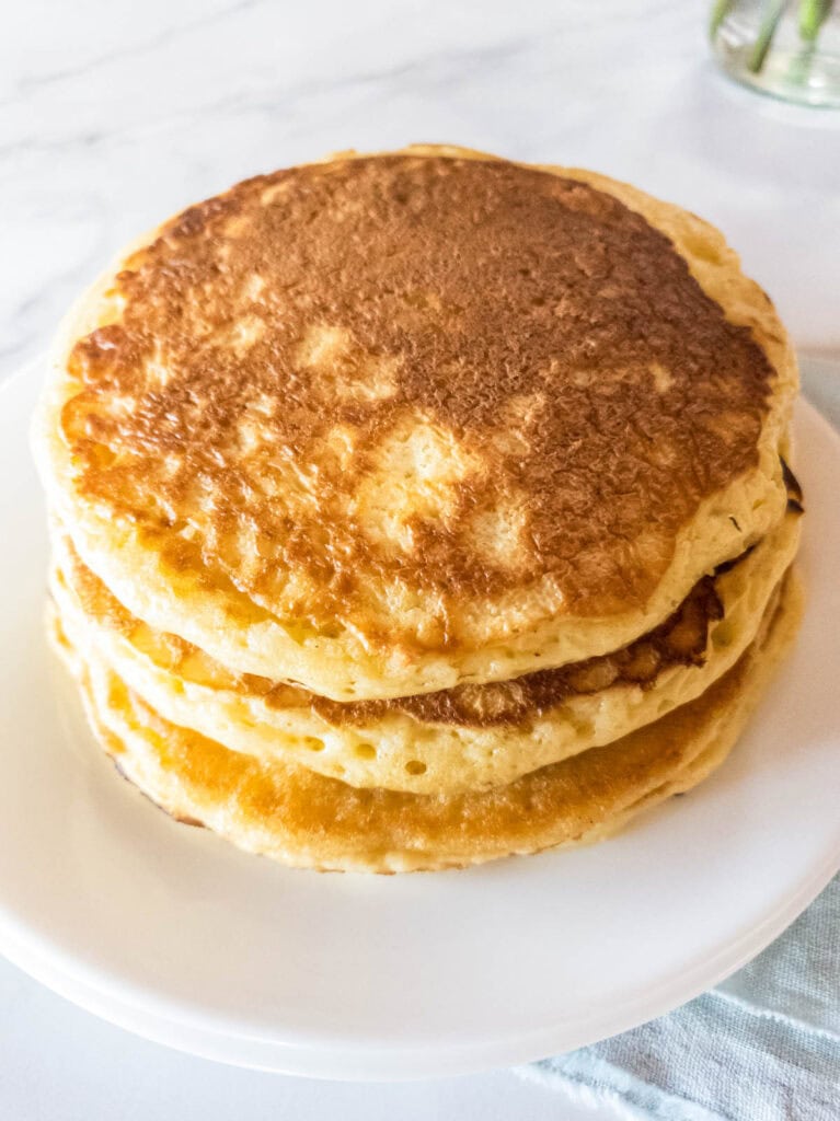 A stack of four golden-brown pancakes on a white plate, set on a light-colored surface.
