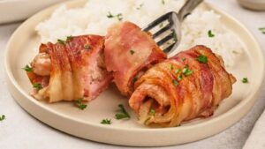 Two bacon-wrapped chicken pieces garnished with parsley on a plate, served with white rice in the background.