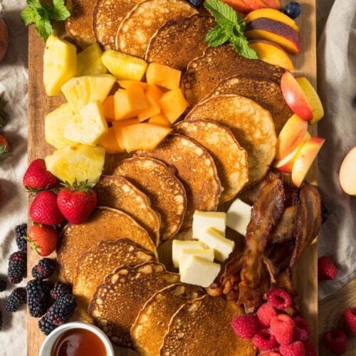 A wooden platter with pancakes, butter, bacon, strawberries, raspberries, blackberries, blueberries, apple slices, pineapple, cantaloupe, and a dish of syrup.