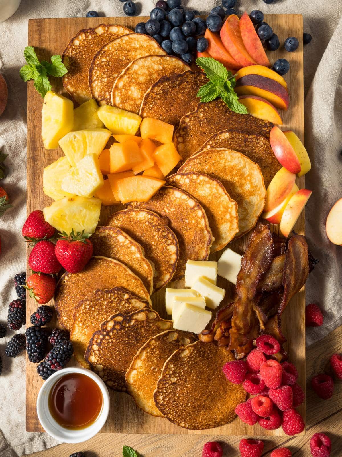 A wooden platter with pancakes, butter, bacon, strawberries, raspberries, blackberries, blueberries, apple slices, pineapple, cantaloupe, and a dish of syrup.