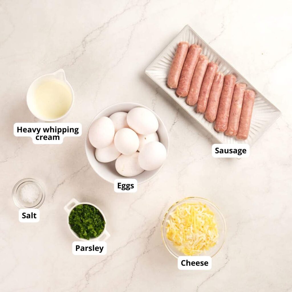 A top-down view of ingredients on a marble counter: heavy whipping cream, eggs, sausage links, salt, chopped parsley, and shredded cheese, each labeled.