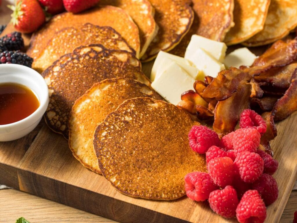 A wooden board with pancakes, bacon, butter, raspberries, blackberries, strawberries, and a small bowl of maple syrup.
