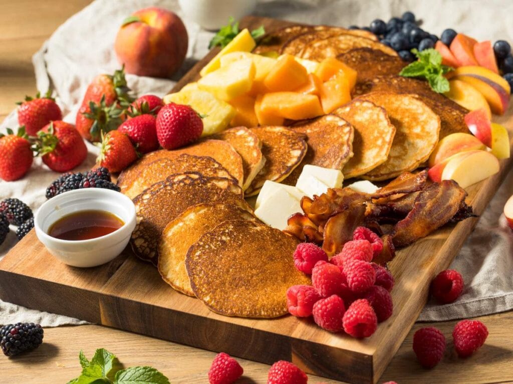 A wooden board with pancakes, assorted fresh fruits, bacon, butter, and a small bowl of syrup arranged neatly for breakfast.