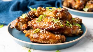 A plate of glazed chicken wings garnished with sesame seeds and sliced green onions, set on a blue plate with a blue cloth in the background.