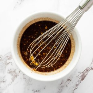 A metal whisk in a white bowl containing a dark, chunky sauce or marinade, placed on a white marble surface.