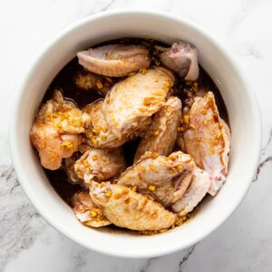 Raw chicken wings marinating in a bowl with a mixture containing chopped garlic and a dark sauce, placed on a white marble surface.